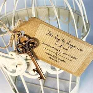 May include: A brown paper tag with the text "The key to happiness is Love (and the ability to open a bottle)" and the names "Jessica & Christopher" and the date "26/08/2023". A vintage-style key with a bottle opener attached is tied to the tag with twine.