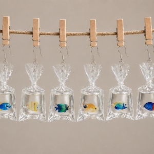 Può includere: Orecchini a forma di pesci in sacchetti di plastica trasparente, appesi a un filo. Ogni sacchetto contiene un pesce colorato, rosso, blu, giallo o arancione. Gli orecchini sono appesi a ganci d'argento e mollette di legno.
