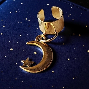 May include: A gold-toned ear cuff featuring a dangling crescent moon charm with a small star cutout. The metal has a polished finish, and the charm is attached to the circular band by a small jump ring, creating a simple, celestial-inspired design.