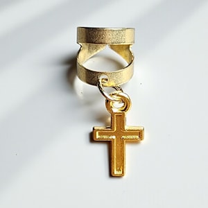 May include: A gold-colored hair accessory featuring a ring with a decorative cutout design and a dangling cross charm. The cross has a simple, classic shape. The accessory is set against a white background.