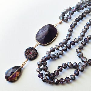 Puede incluir: Un collar largo de cuentas con un colgante. El colgante presenta una piedra grande, ovalada, de color púrpura oscuro con borde dorado, una piedra en forma de lágrima y un colgante circular con un diseño floral. Las cuentas son de color púrpura ahumado.
