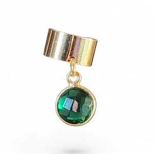May include: A gold-toned ear cuff featuring a smooth, polished band and a dangling circular charm. The charm is set with a faceted, deep emerald green gemstone held in a gold-toned metal bezel, connected to the cuff by a small jump ring.