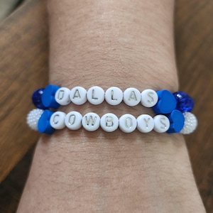 May include: Two beaded bracelets spelling "DALLAS COWBOYS" in white letter beads. The bracelets feature blue and white accent beads. The bracelets are worn on a wrist.