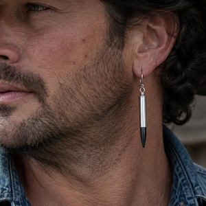 Op de afbeelding: Een man draagt een bruine cowboyhoed en een spijkeroverhemd. Een zilveren en zwarte oorbel in de vorm van een kogel hangt aan zijn oor. De oorbel is een uniek accessoire.