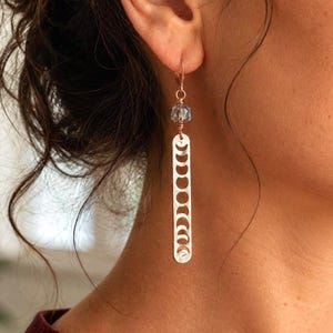 Può includere: Un lungo orecchino pendente caratterizzato da una barra metallica verticale color argento con intagli che raffigurano le fasi lunari. La barra è sospesa a una piccola perla di cristallo azzurro sfaccettata e a un gancio in filo color rame.