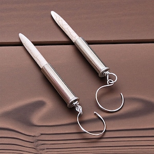 May include: A pair of dangle earrings with white, textured, bullet-shaped pendants and silver-colored metal accents. The earrings have a simple, elegant design with curved ear wires, set against a brown wooden surface.