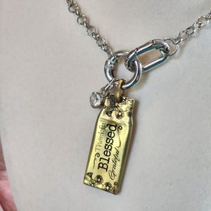 Könnte beinhalten: Eine silberfarbene Kettenhalskette mit einem rechteckigen goldfarbenen Anhänger, der oben und unten mit schwarzen Blumenmustern graviert ist. Der Anhänger trägt die Aufschrift Thankful, Blessed und Grateful und hat einen kleinen klaren Kristall.