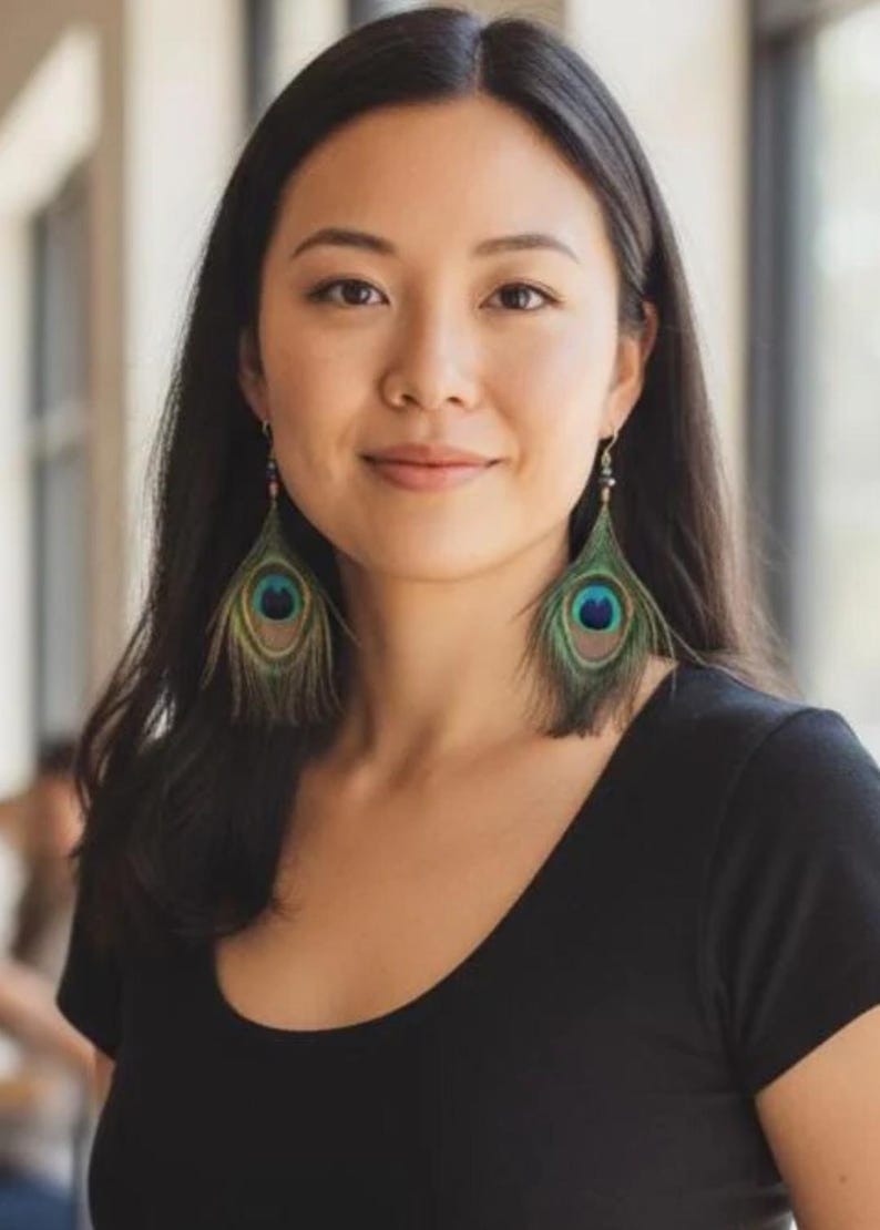 May include: A pair of dangling earrings featuring natural peacock feathers with iridescent green, blue, and brown eye patterns. The feathers are attached to small, multi-colored bead accents at the top, suspended from thin metal hooks.