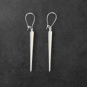 Puede incluir: Un par de pendientes plateados con colgantes largos, cónicos y afilados. Los pendientes tienen un diseño de gancho simple para orejas perforadas. El fondo es una superficie oscura y texturizada.