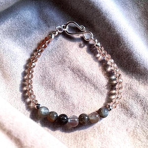 May include: A beaded bracelet featuring a mix of clear and opaque beads in shades of brown, gray, and clear. The bracelet has a silver-colored clasp. The beads are strung together to create a delicate and elegant accessory.