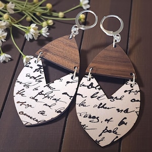 Puede incluir: Pendientes de madera con un diseño único. Los pendientes presentan una parte superior triangular marrón oscuro y una parte inferior en forma de lágrima de color crema con escritura negra. Los ganchos plateados completan el diseño. Un accesorio elegante.