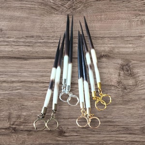 May include: A collection of quill keychains. Each keychain features a natural quill with a black tip and a white base, attached to a metal keyring. The keychains are arranged on a wooden surface, showcasing their unique design and craftsmanship.
