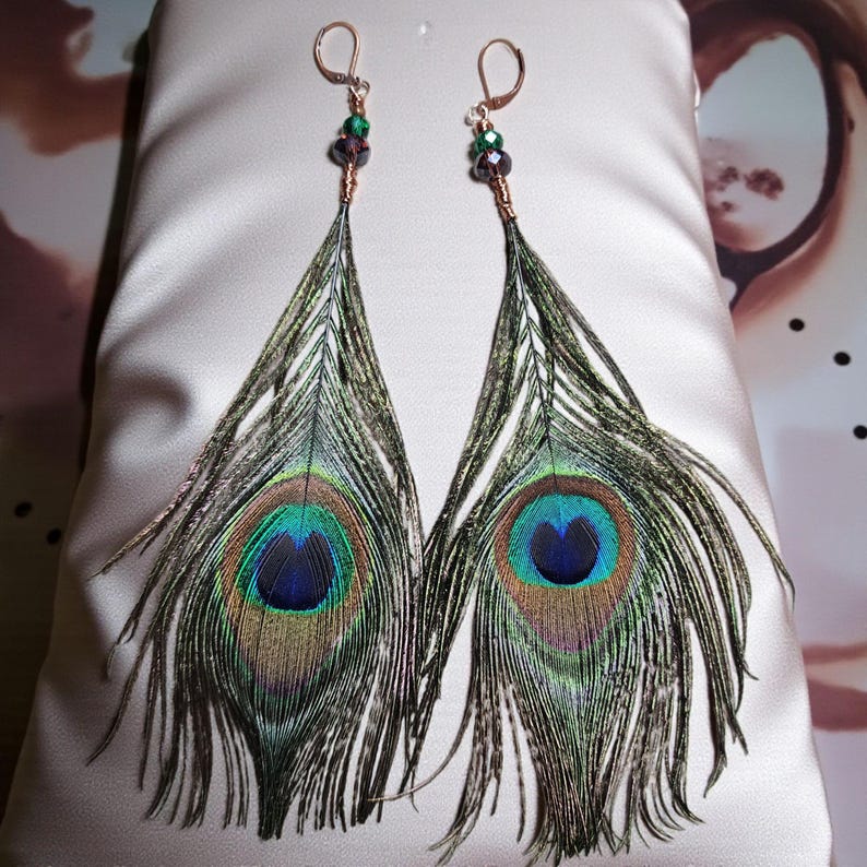 May include: A pair of dangle earrings featuring natural peacock feathers with iridescent blue and green eye patterns. The feathers are attached to copper-toned wire-wrapped accents with small green and dark purple faceted beads and lever-back hooks.