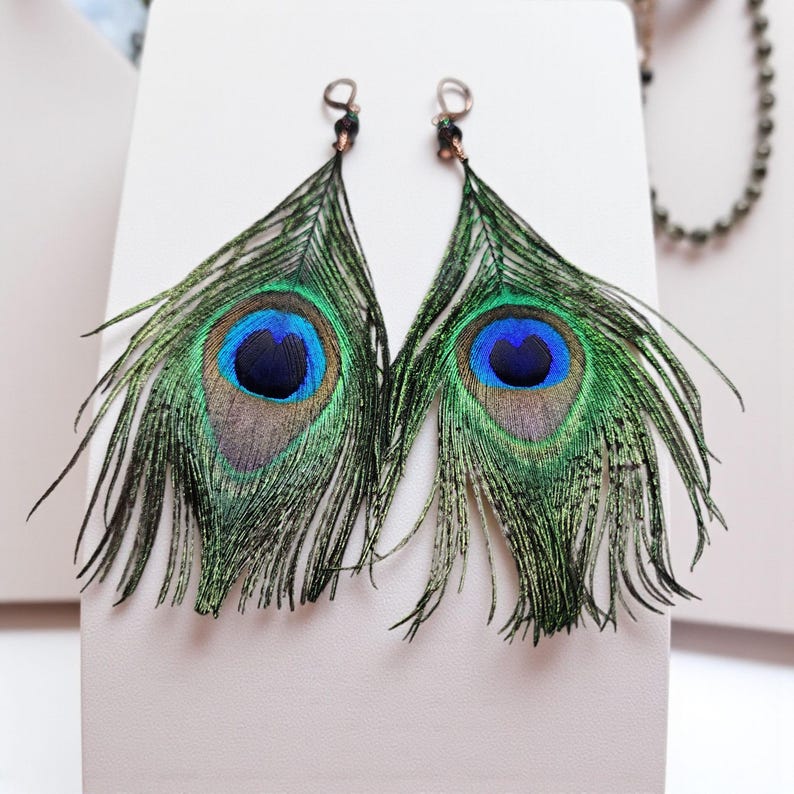 May include: A pair of dangle earrings featuring natural peacock feathers with iridescent green, gold, and blue eye patterns. Each feather is attached to a small metal hook finding with a dark bead accent at the top, creating a long, slender shape.