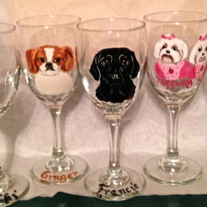May include: A collection of hand-painted wine glasses, each featuring a different pet portrait. The glasses showcase a variety of dog breeds and a cat, with names like "Ginger" and "Bailey" written on the base. The artwork includes detailed depictions of the animals' faces.