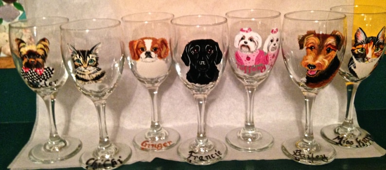 May include: A collection of hand-painted wine glasses, each featuring a different pet portrait. The glasses showcase a variety of dog breeds and a cat, with names like "Ginger" and "Bailey" written on the base. The artwork includes detailed depictions of the animals' faces.