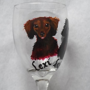 May include: A clear wine glass featuring a hand-painted dachshund dog. The dog is depicted in shades of brown and red with a black outline. The name "Lexi" is written in cursive below the dog.