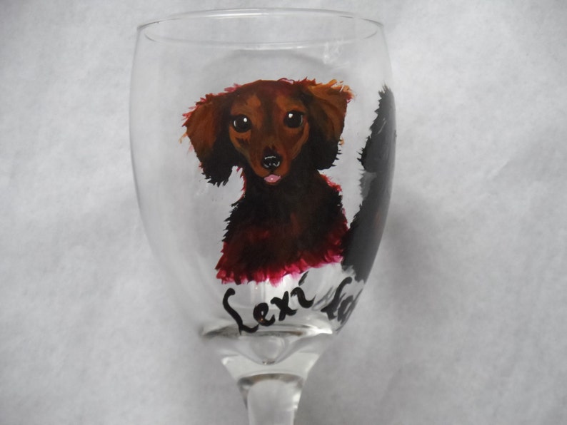 May include: A clear wine glass featuring a hand-painted dachshund dog. The dog is depicted in shades of brown and red with a black outline. The name "Lexi" is written in cursive below the dog.