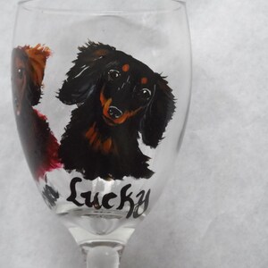 May include: A clear wine glass featuring hand-painted dachshund dog portraits in black, brown, and red. The word "Lucky" is written in black script near the base of the glass. The glass has a long stem.