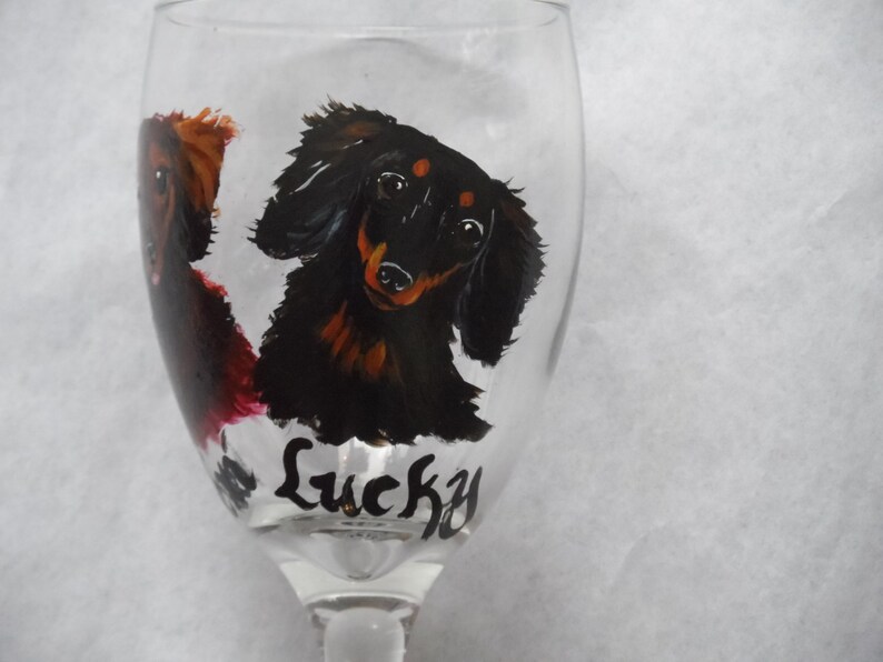 May include: A clear wine glass featuring hand-painted dachshund dog portraits in black, brown, and red. The word "Lucky" is written in black script near the base of the glass. The glass has a long stem.