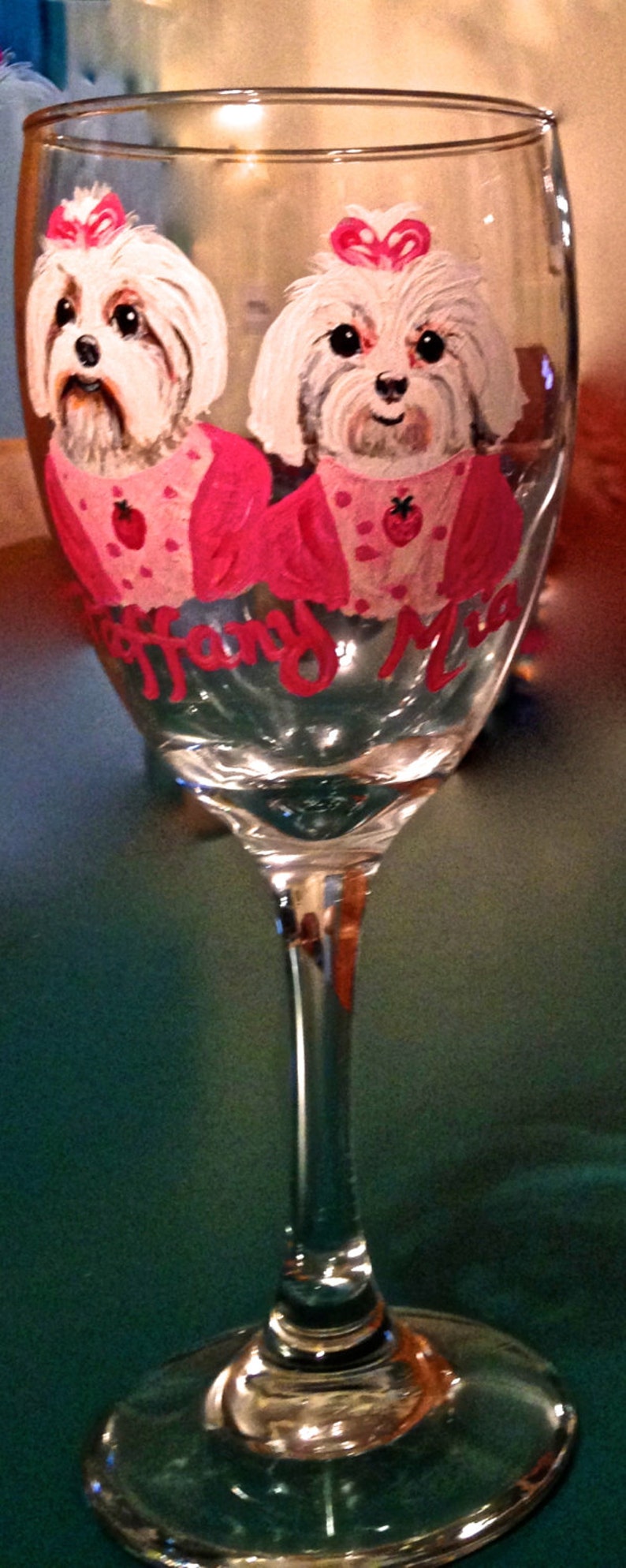 May include: A clear wine glass with a painted design featuring two white dogs with pink bows and strawberry-decorated outfits. The name "Tiffany Mia" is written in red on the glass.