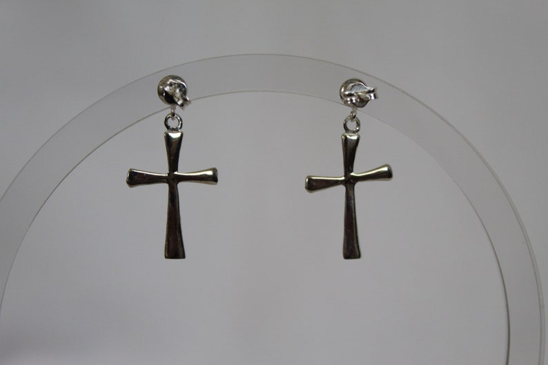 May include: A pair of silver cross dangle earrings. The earrings are on a clear display stand.