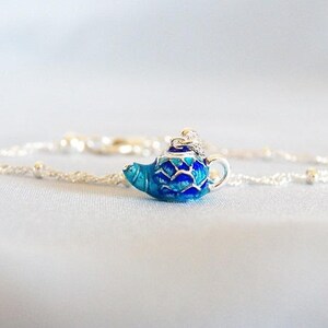 May include: A silver chain necklace with a blue enamel teapot charm. The teapot has a white lid and a silver spout.