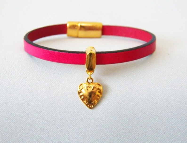Op de afbeelding: Een roze leren armband met een gouden magneetsluiting en een gouden hartbedel.