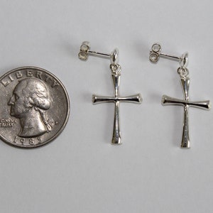 May include: A pair of silver cross earrings with a simple design. The earrings are shown next to a US quarter for size reference.