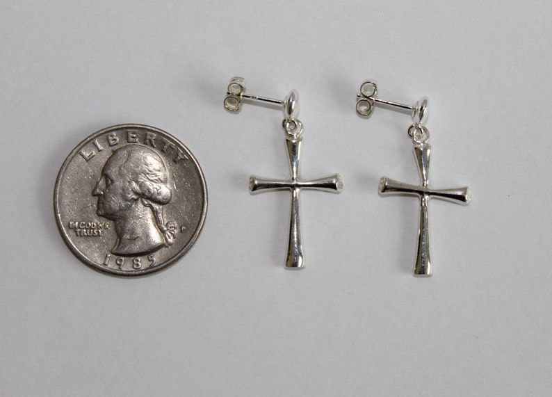 May include: A pair of silver cross earrings with a simple design. The earrings are shown next to a US quarter for size reference.