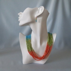May include: A multi-strand necklace with orange, green, and blue beads. The necklace is displayed on a white mannequin bust. Matching earrings are also shown.