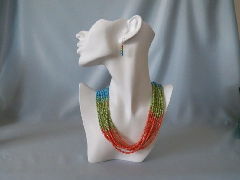 May include: A multi-strand necklace with orange, green, and blue beads. The necklace is displayed on a white mannequin bust. Matching earrings are also shown.