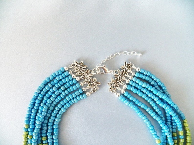 May include: A multi-strand necklace made of turquoise blue and green seed beads with a silver clasp and decorative floral accents.