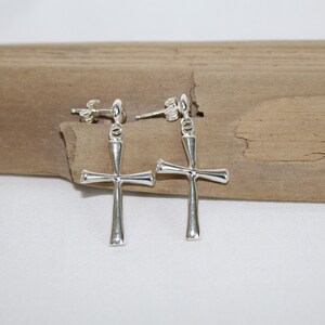 May include: A pair of silver dangle earrings with a cross design. The earrings are on a wooden surface.