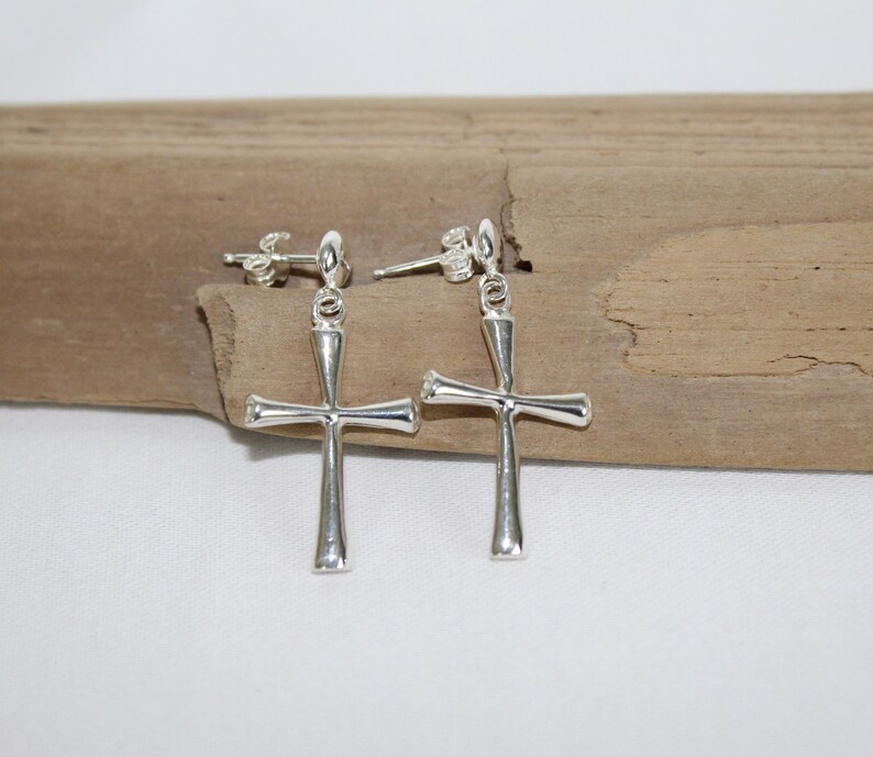 May include: A pair of silver dangle earrings with a cross design. The earrings are on a wooden surface.
