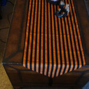 May include: A black and orange striped table runner with a black cauldron and witch's legs on top. The runner is on a wooden table.