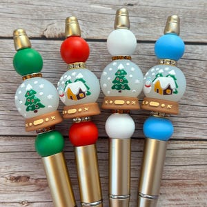 May include: Four festive pens with gold barrels and Christmas-themed toppers. Each pen has a different colour scheme: green, red, white, and blue. The toppers feature a snow globe design with a Christmas tree and a house, perfect for the holiday season.