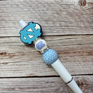 Pu&ograve; includere: Una penna bianca con una parte superiore decorativa azzurra e bianca. La parte superiore presenta un logo azzurro, una perla bianca e azzurra e una perla strutturata azzurra. La penna &egrave; posta su uno sfondo di legno.