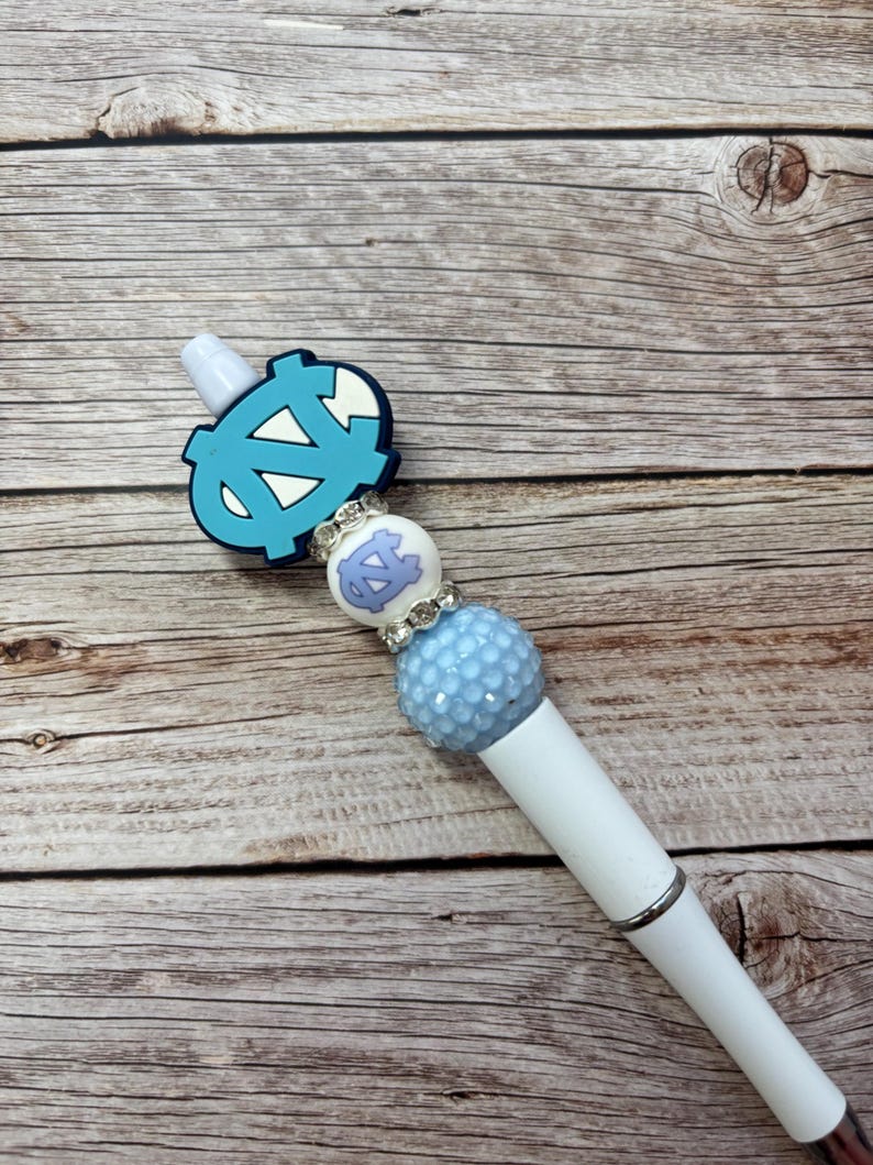 Pu&ograve; includere: Una penna bianca con una parte superiore decorativa azzurra e bianca. La parte superiore presenta un logo azzurro, una perla bianca e azzurra e una perla strutturata azzurra. La penna &egrave; posta su uno sfondo di legno.