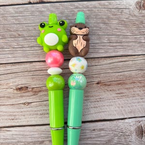Puede incluir: Dos bolígrafos con adornos decorativos. Uno tiene un adorno de rana verde con vientre blanco, el otro tiene un adorno de nutria marrón con sombrero verde. Los bolígrafos tienen una serie de cuentas coloridas y un cuerpo verde o turquesa con detalles plateados.