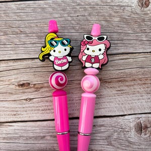 May include: Two pink pens with decorative toppers. One pen features a blonde Barbie character wearing sunglasses, and the other has a pink-haired character with sunglasses. Both pens have a pink and white swirled bead.