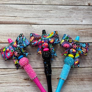 Puede incluir: Tres bolígrafos decorativos con lazos de diseño paisley. Los bolígrafos tienen barriles rosas, negros y azules, cada uno adornado con adornos brillantes. Los lazos presentan patrones vibrantes en tonos azules, rosas, amarillos y verdes.