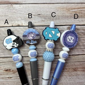 Pu&ograve; includere: Quattro penne con top decorativi con il logo e i colori dei Tar Heels della Carolina del Nord. Le penne hanno vari disegni, tra cui un casco da football e il logo della squadra. Le penne sono nere, grigie, bianche e blu.