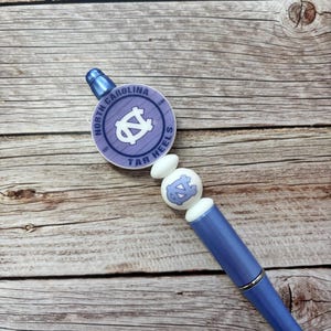 Pu&ograve; includere: Penna azzurra con una parte superiore decorativa con le parole "North Carolina Tar Heels" e il logo della squadra. La penna ha un accento argentato e perline bianche. La penna poggia su una superficie di legno.