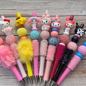 Puede incluir: Una colección de bolígrafos decorativos con adornos de personajes, incluyendo Hello Kitty y My Melody. Los bolígrafos tienen barriles rosas, amarillos y negros, adornados con cuentas y detalles esponjosos. Los bolígrafos se muestran sobre una superficie de madera.