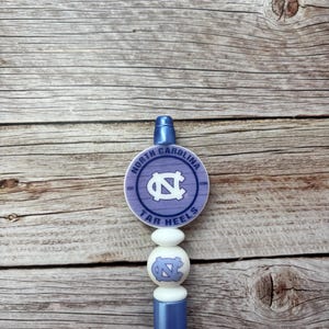 Pu&ograve; includere: Un tappo per bottiglia azzurro con il logo dei Tar Heels della Carolina del Nord. Il tappo ha una parte superiore rotonda, lilla chiaro con il logo e il testo della squadra, e una serie di perline bianche. Il fondo &egrave; di colore azzurro pieno.