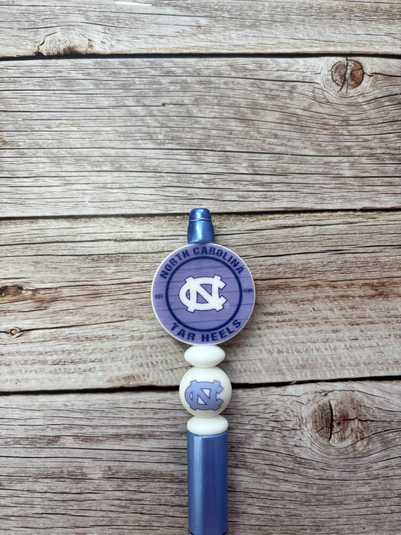 Pu&ograve; includere: Un tappo per bottiglia azzurro con il logo dei Tar Heels della Carolina del Nord. Il tappo ha una parte superiore rotonda, lilla chiaro con il logo e il testo della squadra, e una serie di perline bianche. Il fondo &egrave; di colore azzurro pieno.