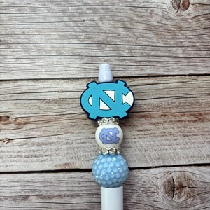 Pu&ograve; includere: Una penna bianca con un design azzurro e bianco. La penna presenta un emblema azzurro con un logo "N" e "C" bianco, una perla bianca con un logo azzurro e una perla azzurra strutturata.