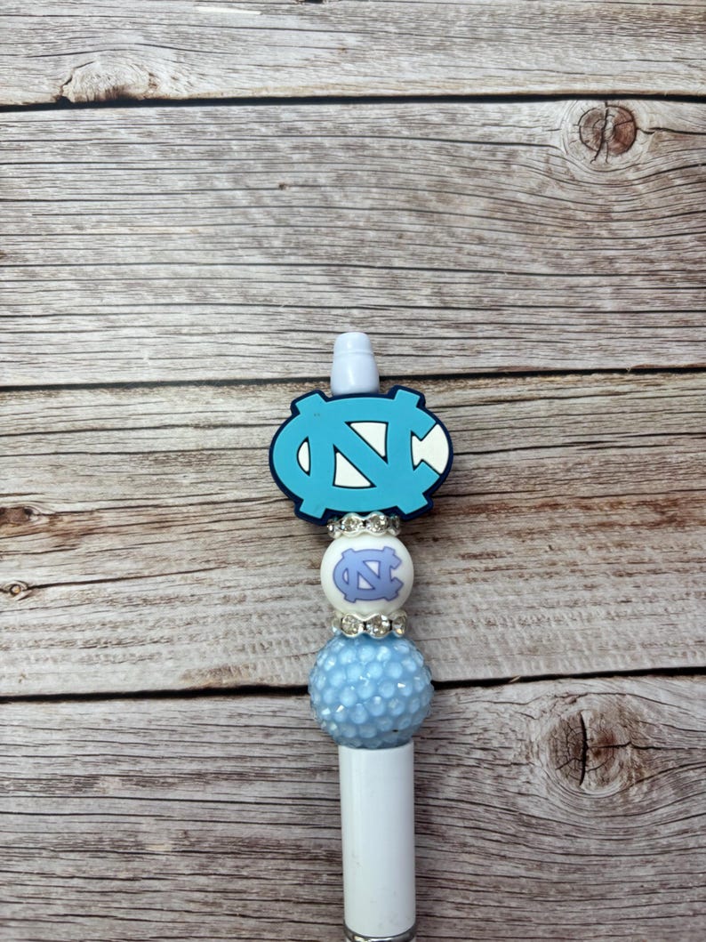 Pu&ograve; includere: Una penna bianca con un design azzurro e bianco. La penna presenta un emblema azzurro con un logo "N" e "C" bianco, una perla bianca con un logo azzurro e una perla azzurra strutturata.