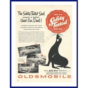 May include: A vintage advertisement for Oldsmobile used cars. The ad features a cartoon seal holding a red circle with the words "Safety Tested Used Car" in white. The text reads "The Safety-Tested Seal means a better Used Car Deal!" and "Every Safety-Tested Used Car is..."